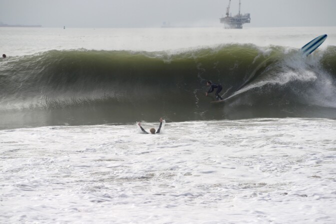 A surfer heads into a big barrel. In front of him in the water, a man holds his hands in the air. On the right, a board shoots up into the air. 