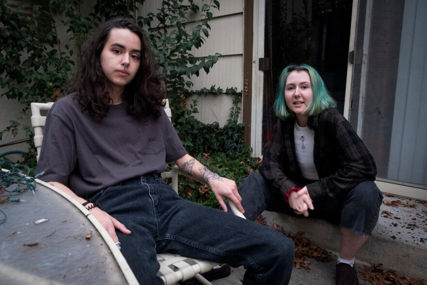 Two young white people sit outside a home. One sits on the step in front of a sliding door, the other sits in a plastic chair at a lawn table. One has blue-green hair, and the other has dark brown hair and a septum piercing.