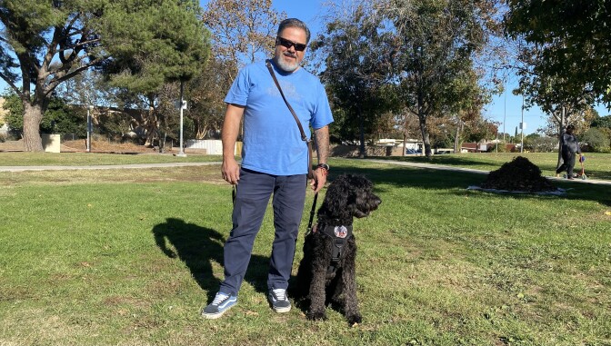 Eddie Aldrete stands on green grass at a park, holding the lead of his dog. The dog is a black Australian Labradoodle named Dakota. 