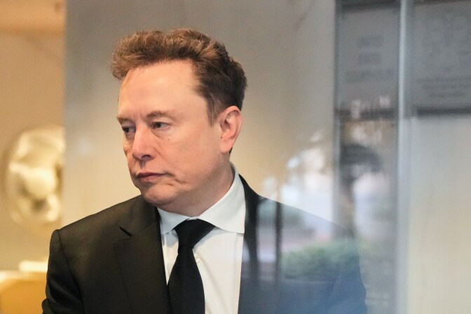 Elon Musk, a man with light skin tone, wearing a black suit and tie, looks out of frame to his right.