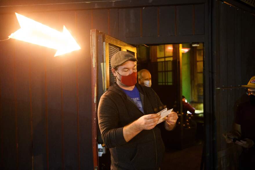 A man wearing a tweed cap and a red cloth face mask stares down at a small white card he's holding in his hand. The scene is darkly lit and a giant, glowing yellow arrow pointing to a doorway behind him.