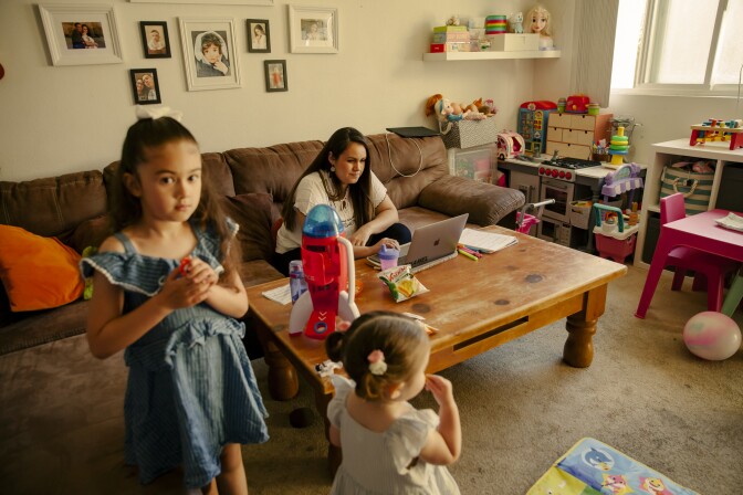 A woman looks at her laptop, while her two daughters play around her in the living room. 