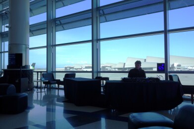A person sits behind a makeshift DJ booth in a public airport lounge area, with tall windows opening up to airport runways behind them.