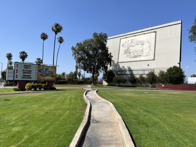 A sign with movie titles and a a screen with the logo "Van Buren Drive-In Theatre" sits between green grass and a concrete walking path 