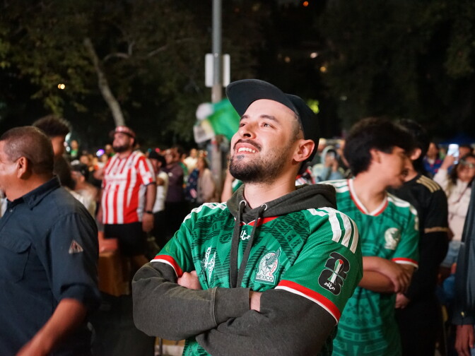 A light skin toned man in a baseball hat and Mexico jersey stands in a crowd, looking up at a screen.
