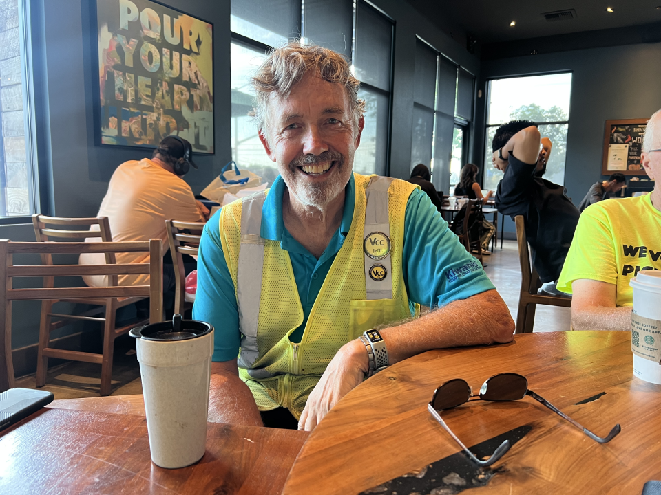 A Volunteers Cleaning Communities member enjoys a refreshing drink with his fellow volunteers after a clean-up session. His sunglasses are on the table. He wears a real shirt with his yellow vest over it. He smiles at the camera with his elbow on the table.