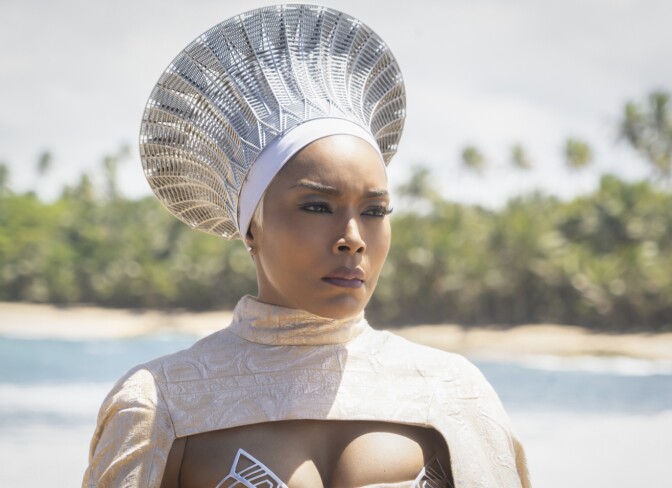 A dark-skinned woman, Angele Bassett, is in the foreground, wearing an African style futuristic headress and elegant robe. There is a beach with palm trees in soft focus in the background.