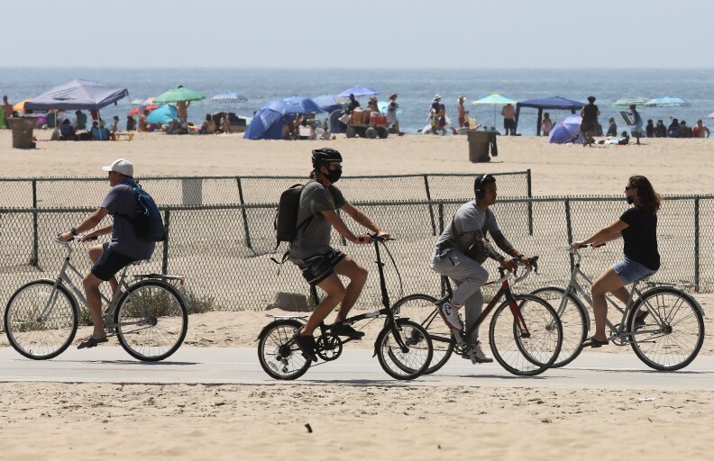 People on bikes ride on a cement path on a beach. Beachgoers with umbrellas and tents sit by the ocean in the background.
