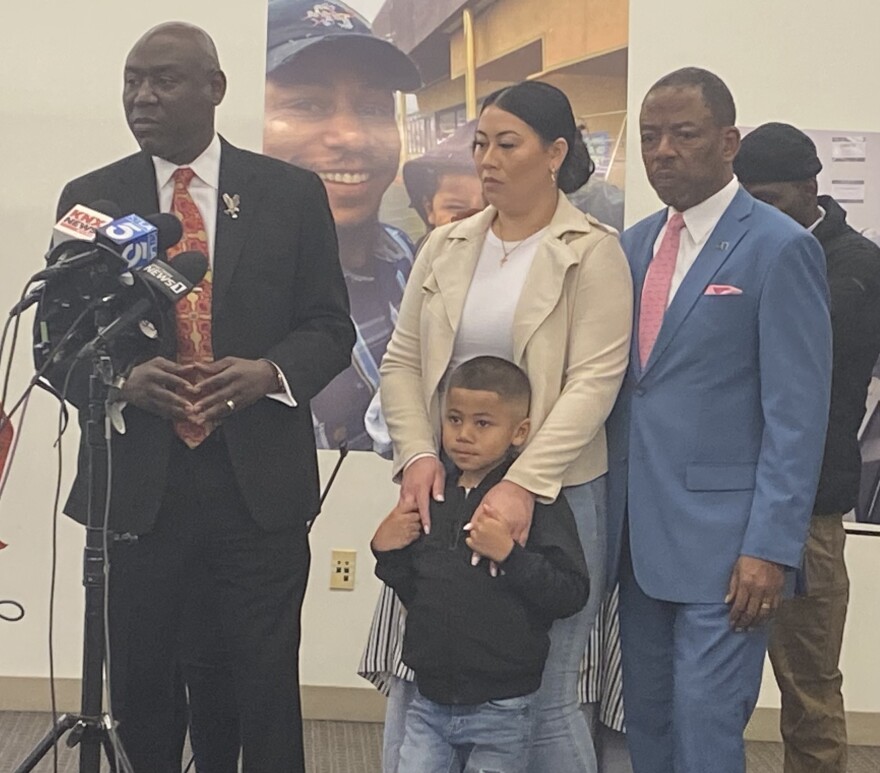 Attorney Ben Crump, wearing suit and tie, speaks to reporters in front of an amplified photo of Keenan Anderson, who smiles wearing a cap. Crump is joined by Gabrielle Hansell, the mother of Anderson's five-year-old son.