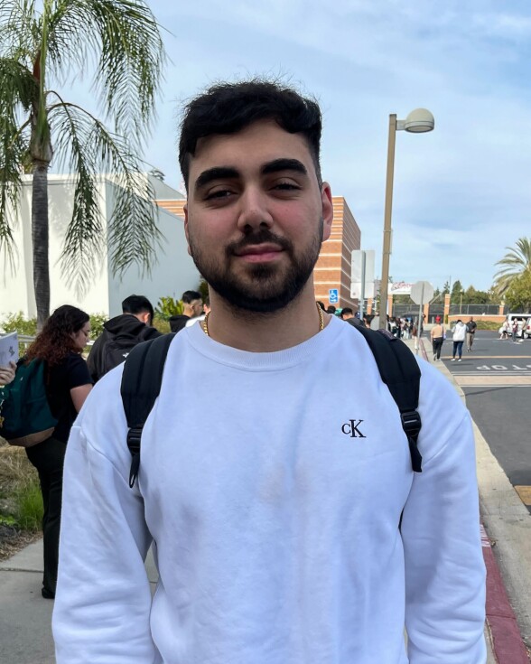A man with short dark hair and goatee wearing a white sweater stands on a sidewalk. 