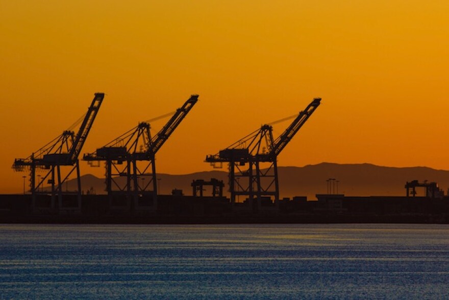 Three large machines are shown on the horizon of the Port of Long Beach. A vibrant orange sunset is in the background, and the blue water is in the foreground.