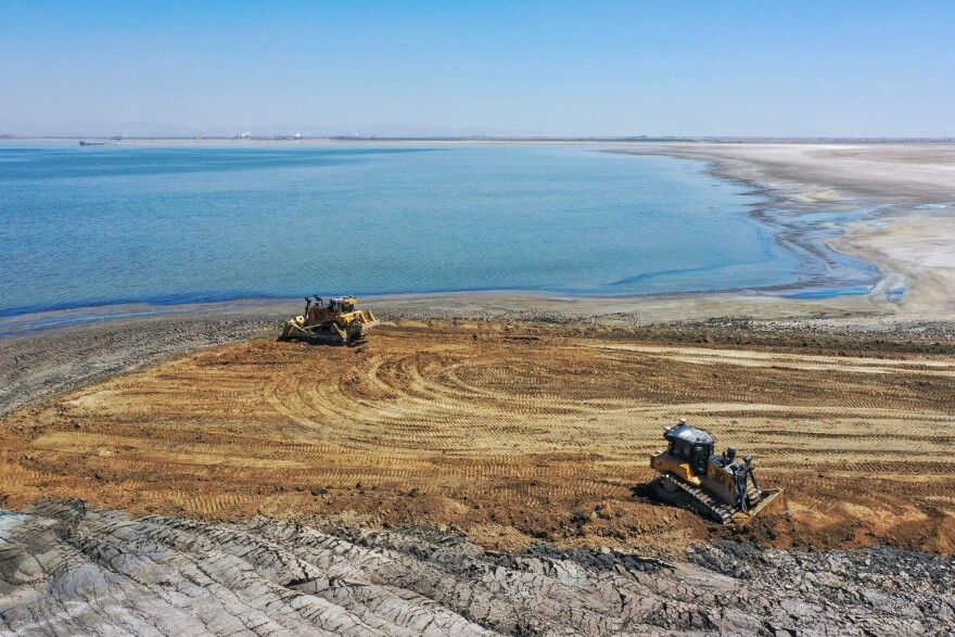 Two bulldozers are pictured pushing dirt on the shore of lake.