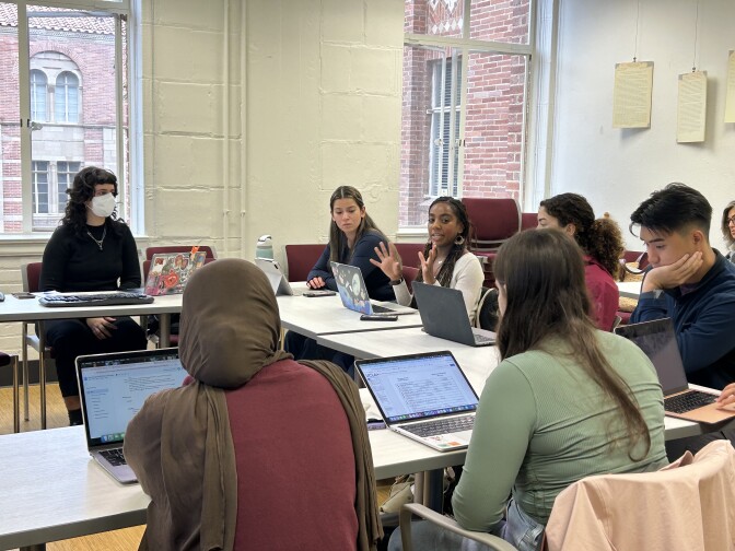 Eight people sit in a classroom with large, open windows that look out into neighboring brick buildings. The students pause from taking notes on their laptops to listen to a classmate with medium skin tone, long hair and hoop earrings in the center of the image. 