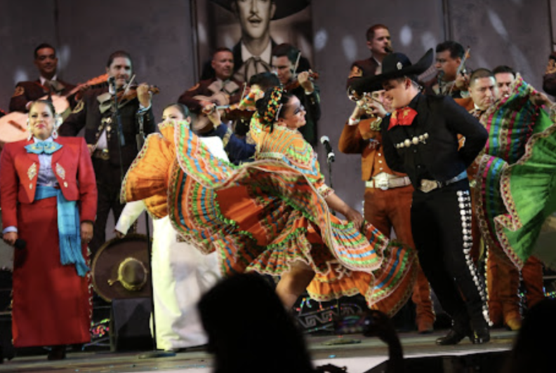Dancers in colorful Mexican ballet folklorico attire, and mariachi musicans in black Mexican charro suits, perform on a stage.