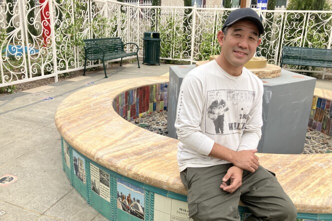 A man in a baseball cap, white graphic t-shirt and olive green pants sits at a fountain that has a yellow stone top and blue, colorful tiles depicting various images.