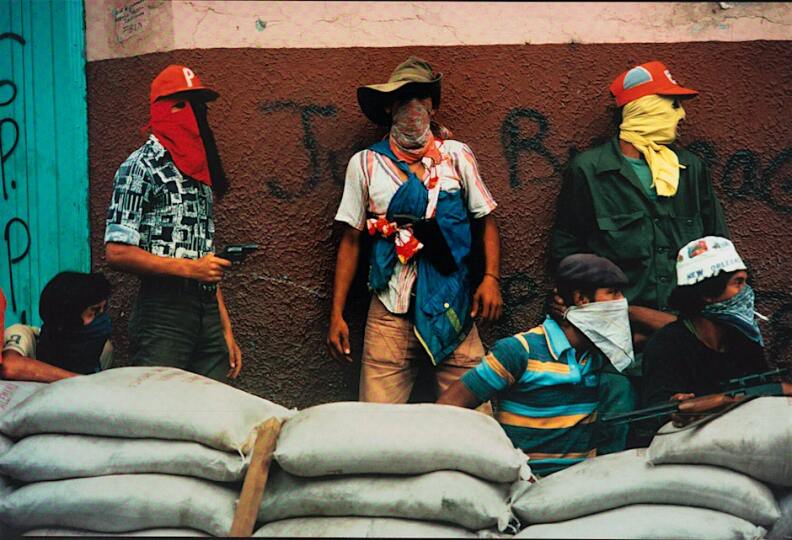 A photograph of six men, all wearing face coverings and some holding guns, braced behind sandbags and seemingly in the middle of a battle.
