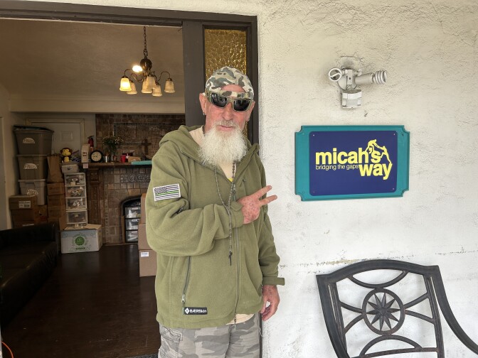 A man with a long, white beard, wearing an army green sweatshirt, sunglasses and a backwards camo-print baseball hat, stands in front of an open doorway with two fingers pointing toward an adjacent sign that reads "micah's way bridging the gaps."