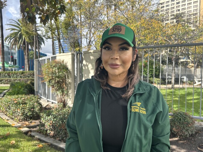 A woman with medium skin tone and long, gold and brown hair smiles gently for a photograph. She is clad in a green jacket and baseball cap, both labeled “AFSCME 3299.”