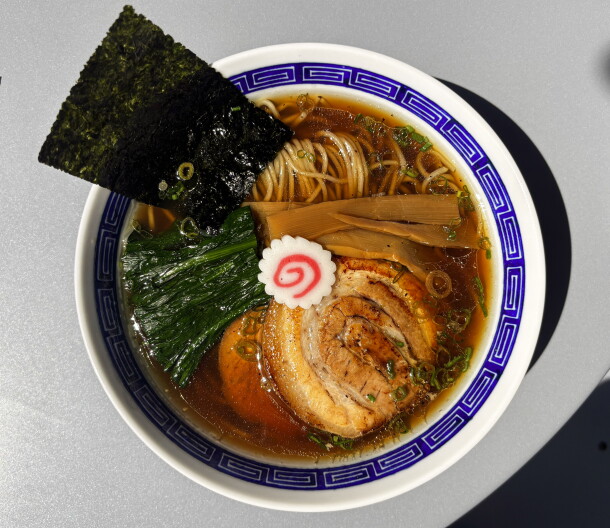 An overhead shot of a blue-and-white ramen bowl filled with brown broth, noodles, sliced pork chashu, a triangle of nori, spinach and a pink-and-white fish cake, set in direct sunlight.