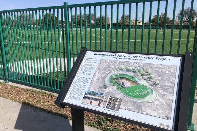 An educational sign shows a diagram of a soccer field with a filtration system beneath it. In the background is a green fence surrounding a turf soccer field. 