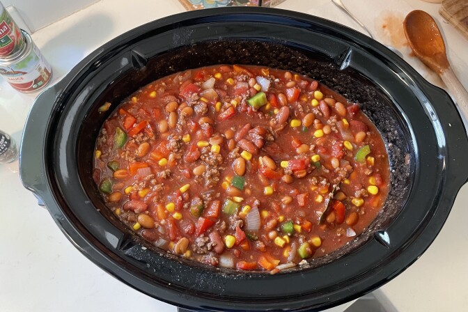 An overhead image of a large black oval-shaped ceramic pot. The dish is filled with a red liquid and contains bits of brown cooked meat mixed with cut-up green peppers, red tomatoes, transparent white onions, yellow kernels of corn, and red kidney beans. The pot sits on a white counter space. To the left are two canned foods stacked on each other, and to the right is a wooden spoon with the top wet with a red liquid similar to the one in the pot.