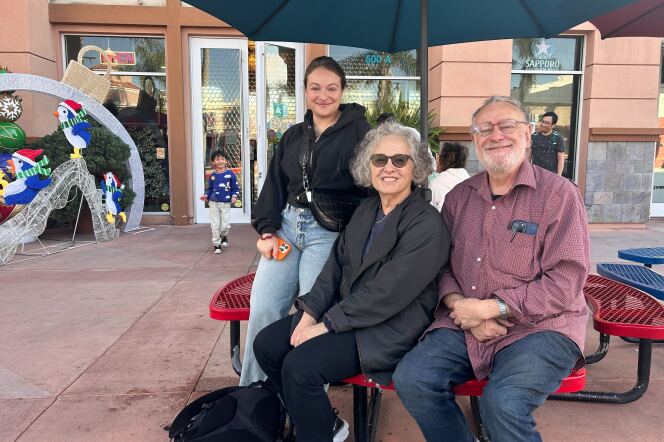 A young woman wearing blue jeans and a black jacket poses and smiles with an older woman in a black jacket and an older man in a red long-sleeve button down shirt. They are sitting at a picnic table in front of a restaurant that has Christmas decorations in the corner. 
