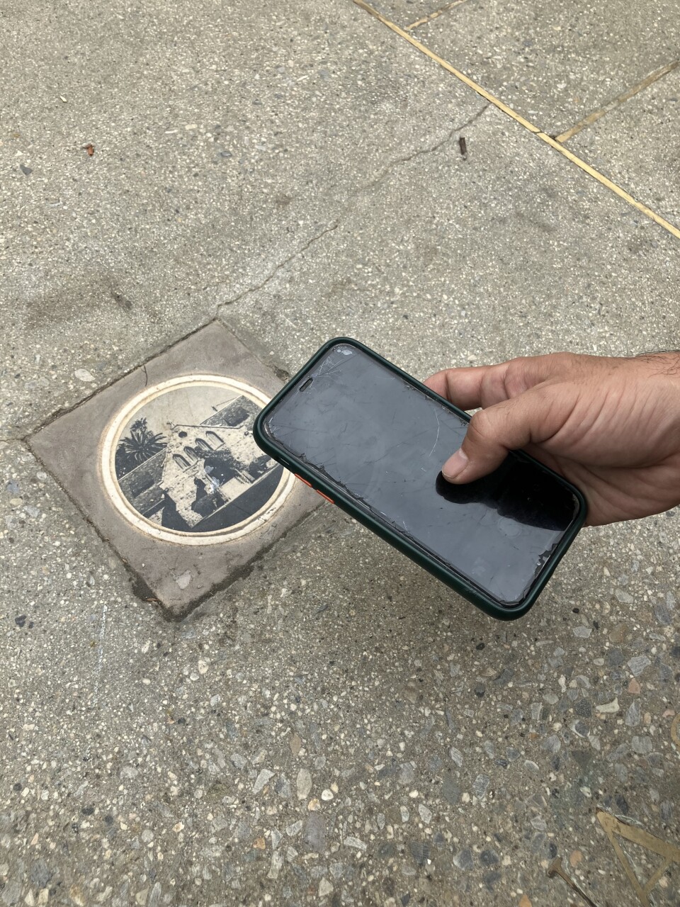 A man's hand holds a cell phone above a tile embedded in concrete. The tile depicts an image of a peaked-roof building.