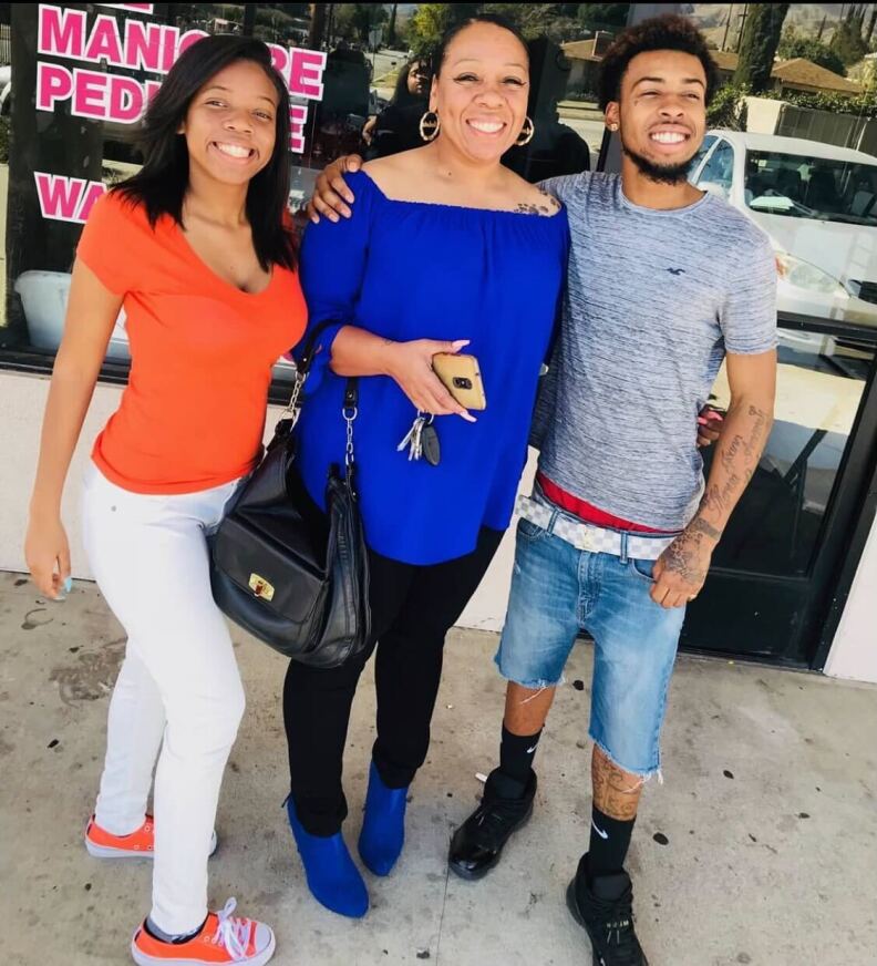A Black woman in a bright blue shirt holds a cell phone while smiling. She's flanked on the left by a young Black man in a gray T-shirt and jean shorts with tattoos on his arm and leg, and young Black woman to her right wearing a bright orange top and sneakers.
