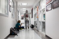 Two students in a bright school hallway: one sits on the floor against the wall eating a snack beside backpacks and a lunch bag, while another in a gray hooded sweatshirt walks away from the camera.