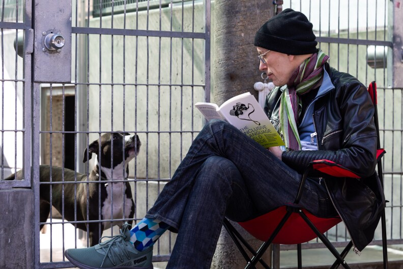 A man with light skin wearing jeans, a black beanie, glasses, and layered sweaters sits in a portable red chair next to a metal dog kennel. He's holding an open book while looking at a black and white pitbull inside the kennel.