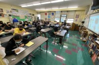 Students seated in a first period class at Narbonne High School, an L.A. Unified School District campus in Carson. Students are seated at long tables, writing in notebooks and binders and viewing images on a Smartboard at the front of the classroom.