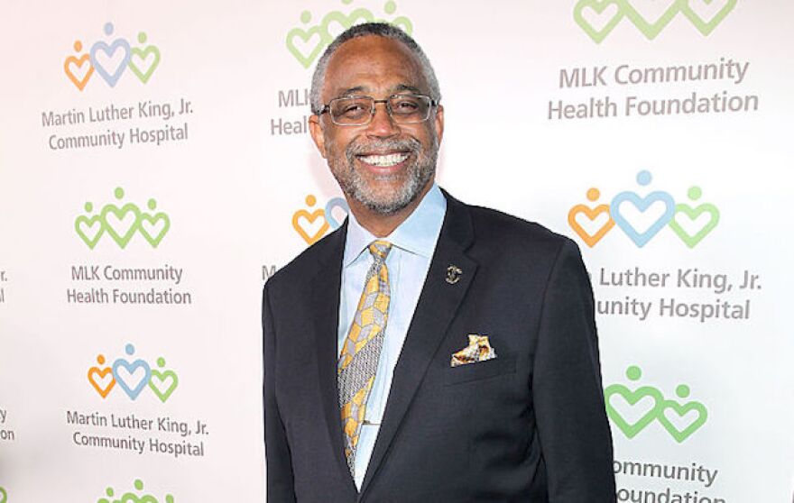 A Black man in a suit with glasses and gray hair smiles against a backdrop covered with logos from the MLK Community Health Foundation