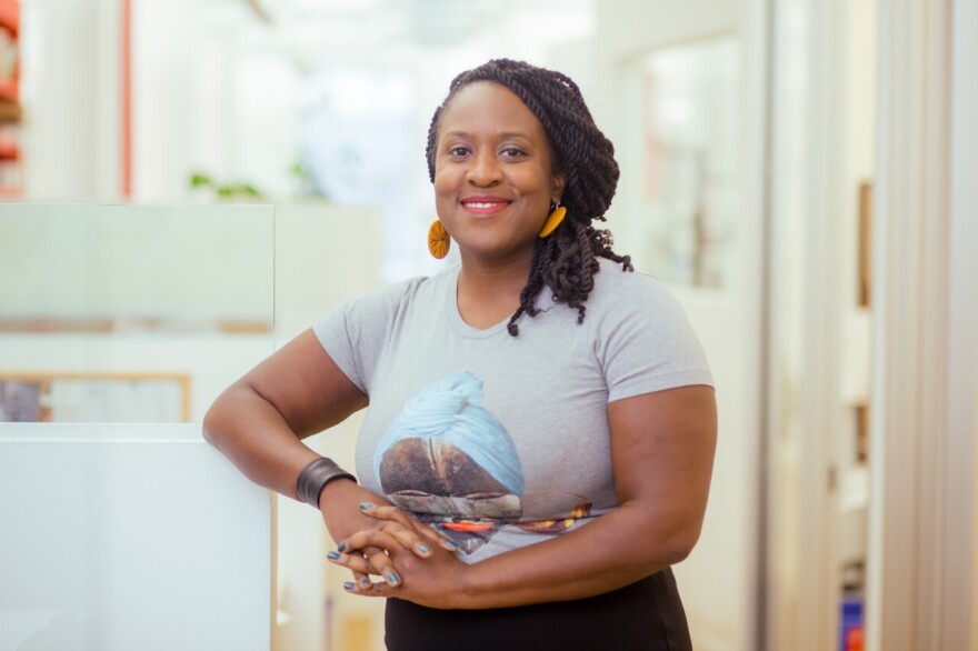 Priscilla Ocen stands facing the camera, her hair swept down the left side of her head, wearing a grey t-shirt and large round orange earrings, with a dark band on her right wrist. She is leaning her right elbow on the top of an office cubicle.