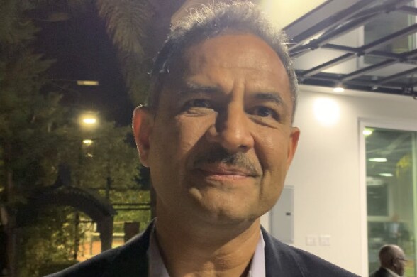 A Pakistani American man wearing a moustache and a suit looks into the camera. Behind him is the night sky and an open-air venue.