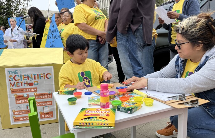 A 4-year-old boy with light brown skin plays with playdough at a children's table set up as part of a rally. People hold signs, chanting behind him. 