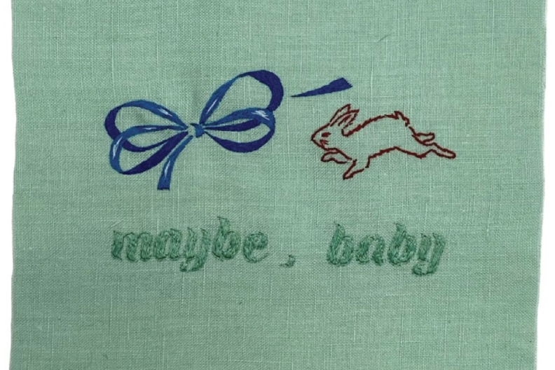 A green tapestry with the words "maybe, baby" stitched into it in a dark green. There's also a blue blow and rabbit. 