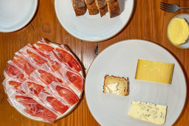 A series of white ceramic plates are arranged on a medium-dark wooden tabletop. In the bottom left corner, overlapping slices of red and white cured ham cover one plate entirely. To the right, three small cheese slices display a range of colors: pale yellow, light mustard brown, and white, which is speckled with small black dots. Above this plate is a small dish containing a portion of yellow butter. At the very top, four slices of brown sourdough bread are positioned closely together on their sides.