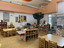 The interior of a classroom for young children, with wooden tables and chairs neatly arranged around cubbies filled with toys.
