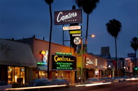 Canter's Deli's neon sign glows above Fairfax Avenue at dusk, reading "Restaurant," "Open 24 Hours," and "Bakery," with palm trees and passing car lights in the foreground.