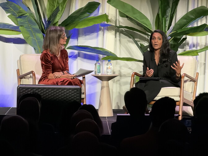 Two women sit in chairs on a stage, one listening to the other.