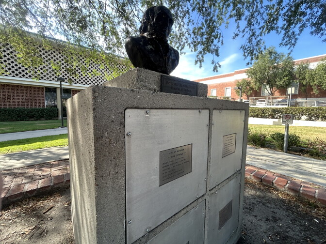 A cement monument with four square compartments topped by a black bust of a man. The monument is outside on a sunny day between two buildings and patches of grass.