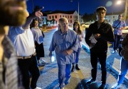 A man walks at night in a blue outfit surrounded by other people, along a road with buildings in the background, as someone points in the direction the man is looking.