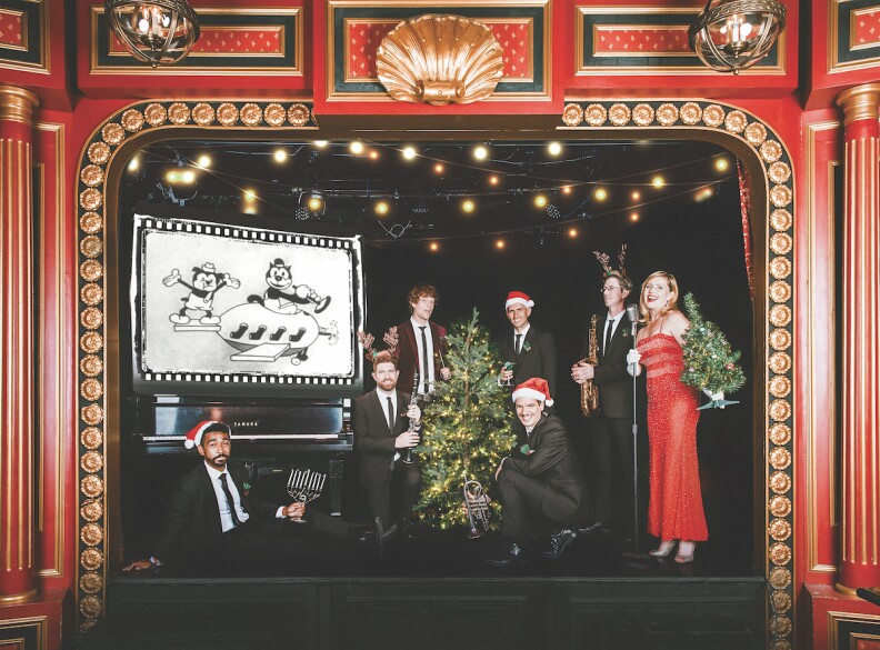 Six men in suits and holiday hats stand and sit on a stage next to a woman holding a small Christmas tree.