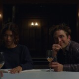 A female presenting person and a male presenting person sitting side by side at a table, across from two other figures who are not visible. Wine glasses are on the table.