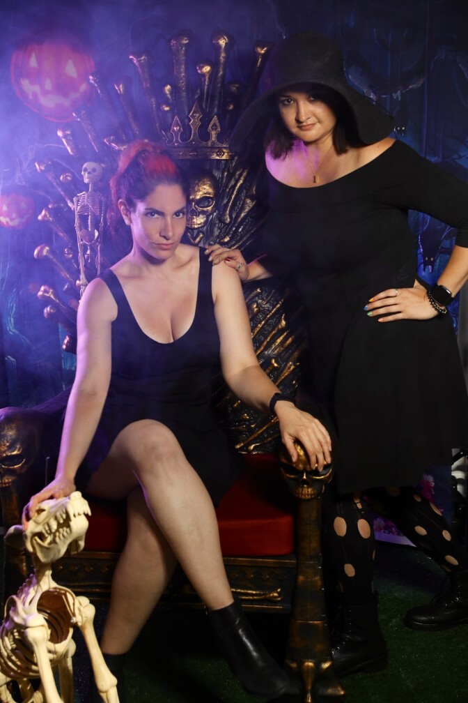 A person wearing a dress and crossing their legs in a throne with hair including bright red dyed highlights, next to a woman in a dress and a hood. There are spooky decorations including pumpkin designs and animal skulls, along with spooky lighting and fog.
