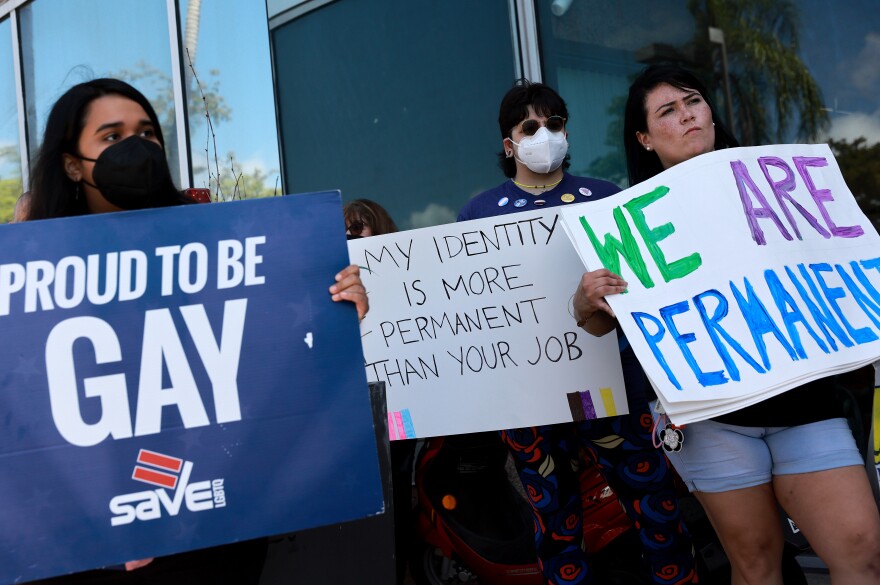 People, two masked, one unmasked, carry signs that read: "Proud to be Gay" "My identity is more permanent than your job" and "We are permanent."