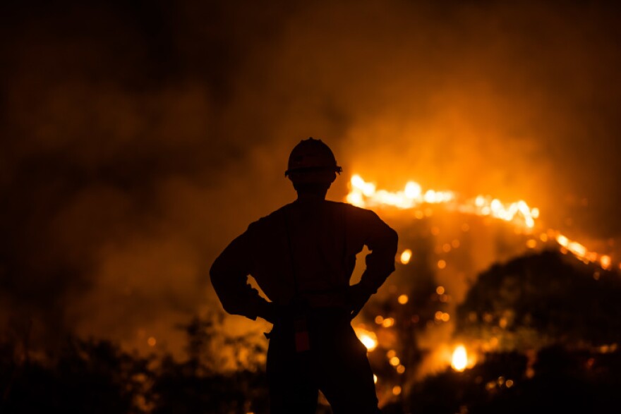 A person is silhouetted against red flames of a fire.