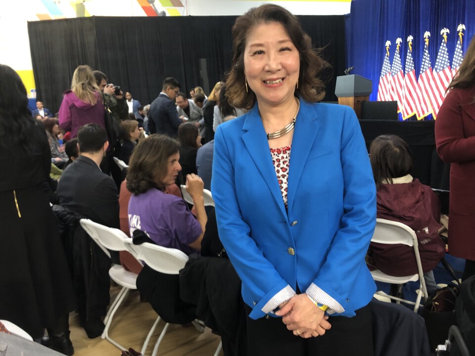 An Asian woman wearing a bright blue blazer over a floral top and a three-ring silver necklace poses in a room where hundreds of people are seated in folding chairs set in front of a stage with a podium and several American flags.