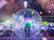 Fireworks and confetti light the stage as a performer stands inside a big plastic bottle.