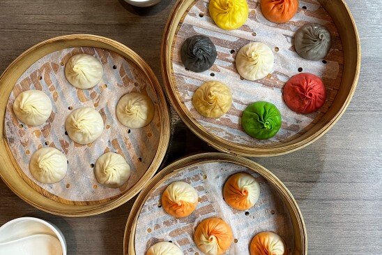 A photograph of three sets of dumplings in bamboo steamer baskets, each a different color, sitting on a wooden table. The bottom group of dumplings is orange and white. The center group is white. The upper right basket contains dumplings that are green, red, grey, back, yellow, orange and white. 